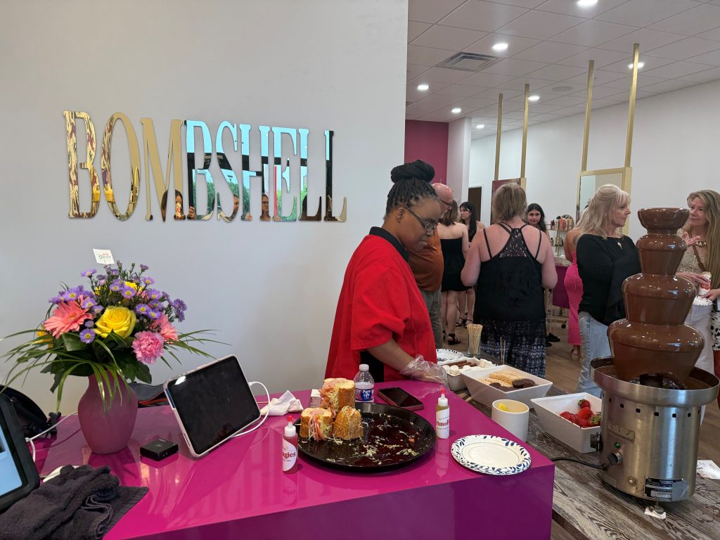 Photo of ribbon cutting for Bombshell Hair and Beauty Studio's 563 New Scotland location, Bombshell is written on the wall, there is a chocolate fountain, and people are gathered socializing in the reception area. 