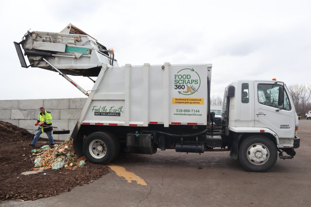 Photo: A white truck with the logo "FOOD SCRAPS 360" on the side is shown dumping organic waste onto the ground. The truck has the slogan "Feed the Earth, NOT THE LANDFILL!" written on the side. The contact information includes a phone number, 518-888-7144, and a website, www.foodscraps360.com. A man wearing a bright yellow-green safety jacket, jeans, and gloves is standing near the pile of organic waste, which includes food scraps in green bags and loose items. The background shows a concrete block wall and some trees under an overcast sky. The truck's rear compartment is raised to unload the contents. The setting is the town of bethlehem's compost facility.