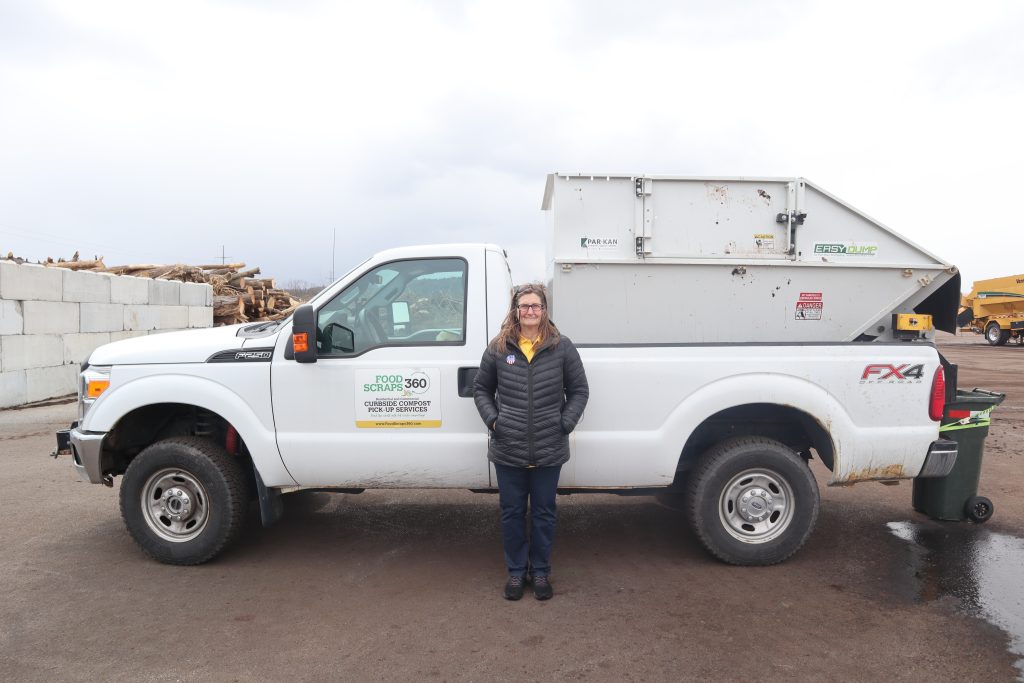 Photo: A woman (Diana) stands in front of a white pickup truck with a large white container attached to the back. She has long brown hair, wears glasses, a black puffer jacket, a yellow shirt underneath, dark pants, and black shoes. The truck has a logo on the door that reads "FOOD SCRAPS 360" with additional text about curbside compost pick-up services and a website address. The container on the truck has labels including "PARKAN" and "EASY DUMP." The truck is parked on a paved surface with a stack of cut logs behind a concrete block wall on the left side of the image. The sky is overcast. The woman is smiling slightly and has her hands in her jacket pockets.