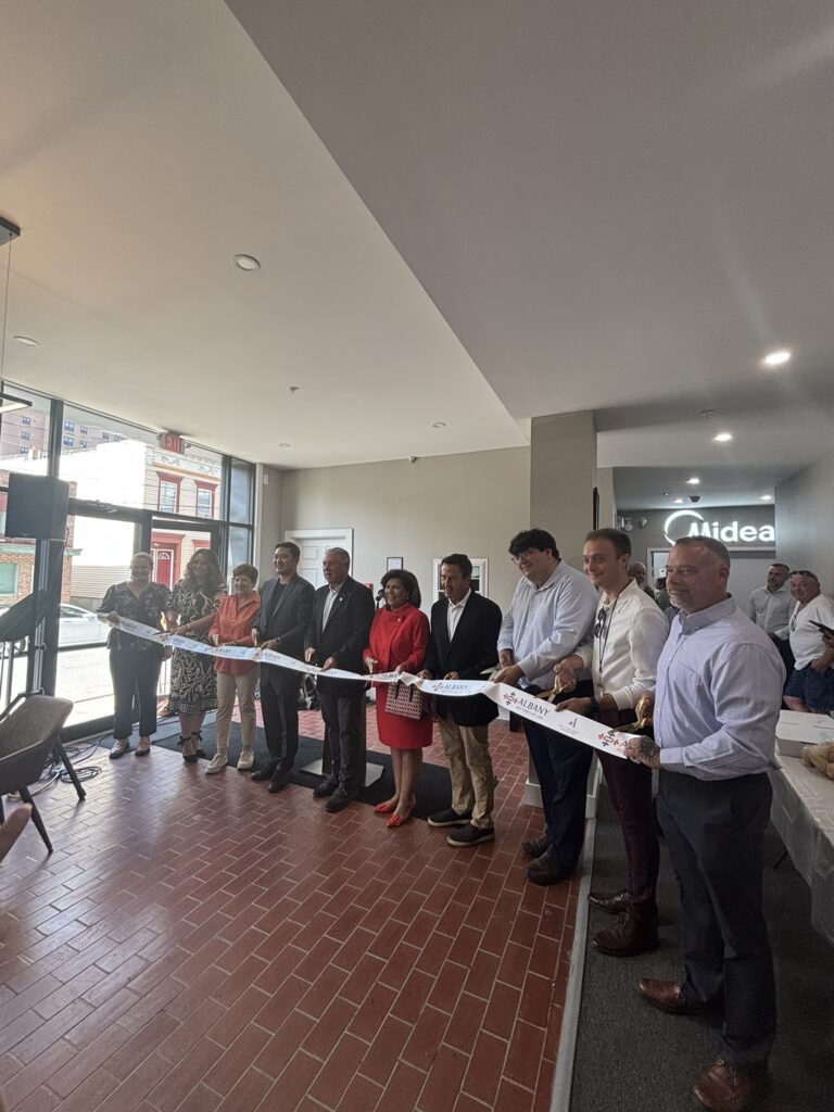 A group of nine people, dressed in business attire, stand in a line indoors holding a white ribbon with “Albany” and a star logo printed on it. Each person holds a large pair of gold scissors, smiling and posing for a ribbon-cutting ceremony, with natural light coming through large windows in the background.